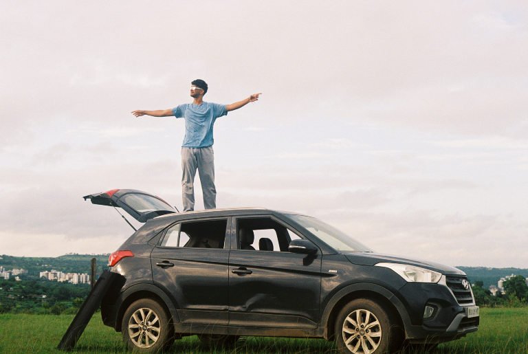 A person standing on the roof of a parked car with both arms outstretched, fields and hills behind