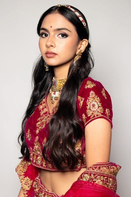 Bridal makeup: a model in a red and gold embroidered lehenga with a maang tikka, pale backdrop