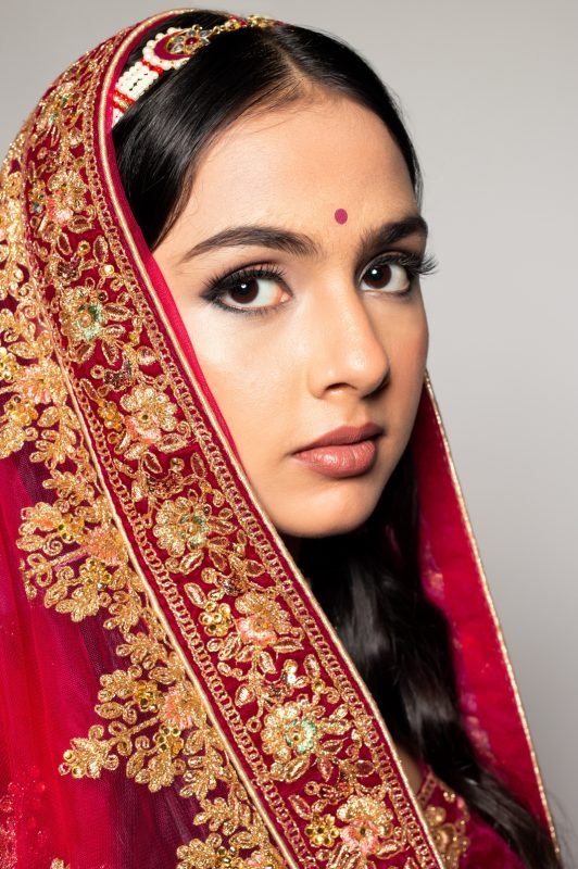 Close bridal portrait: a red and gold embroidered dupatta framing the face, defined eye makeup and a bindi